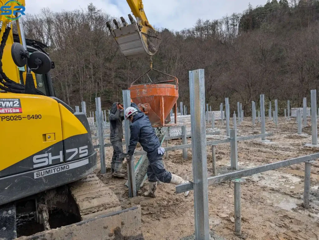 Workers pouring concrete for SR Solar ground mounting system pillars using an excavator on a mountain solar project site.