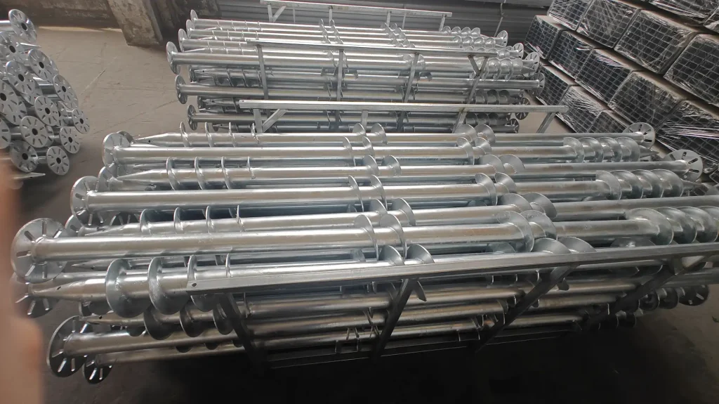 SR Solar double wide-blade helical ground screw foundation anchors stacked neatly in factory steel frames